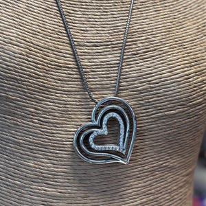 Pode incluir: Colar prateado com um pendente de cora&ccedil;&atilde;o triplo. O pendente tem um design em camadas, com o cora&ccedil;&atilde;o mais interno adornado com pequenos cristais brilhantes. O colar &eacute; exibido em um manequim.