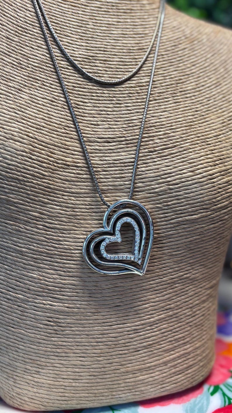 Pode incluir: Colar prateado com um pendente de cora&ccedil;&atilde;o triplo. O pendente tem um design em camadas, com o cora&ccedil;&atilde;o mais interno adornado com pequenos cristais brilhantes. O colar &eacute; exibido em um manequim.