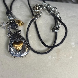 May include: Silver-toned pendant necklace with a teardrop-shaped charm featuring a gold heart. The necklace has a black cord and decorative silver accents. A small gold leaf and crystal beads are also present.