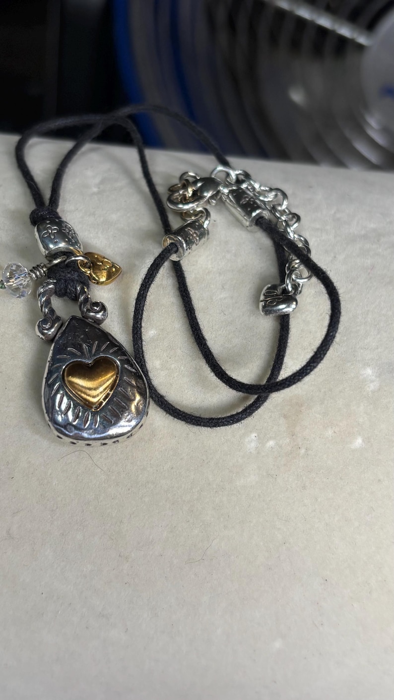 May include: Silver-toned pendant necklace with a teardrop-shaped charm featuring a gold heart. The necklace has a black cord and decorative silver accents. A small gold leaf and crystal beads are also present.