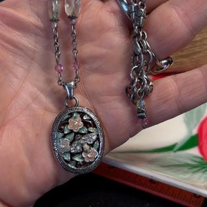 Puede incluir: Collar plateado con un colgante ovalado con un dise&ntilde;o floral y de abeja. El colgante tiene un fondo de esmalte verde claro y rosa. El collar tiene una cadena delicada con detalles de cuentas rosas y transparentes.