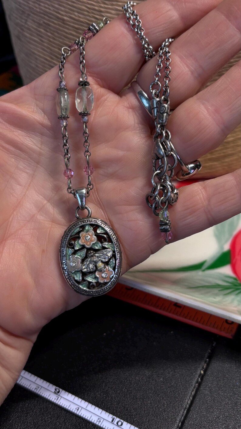 Puede incluir: Collar plateado con un colgante ovalado con un dise&ntilde;o floral y de abeja. El colgante tiene un fondo de esmalte verde claro y rosa. El collar tiene una cadena delicada con detalles de cuentas rosas y transparentes.