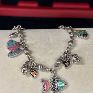 Pu&ograve; includere: Bracciale con ciondoli in argento con ciondoli in smalto colorato. Il bracciale presenta ciondoli a forma di pesce, cuori, farfalle e campane. I ciondoli sono dipinti con colori vivaci tra cui rosa, verde, blu e arancione.