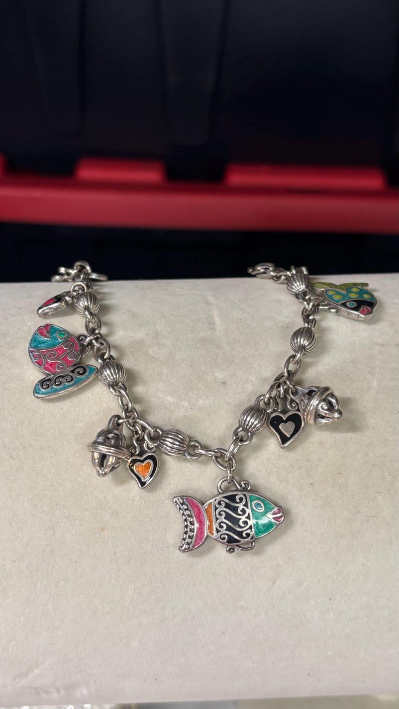 Pu&ograve; includere: Bracciale con ciondoli in argento con ciondoli in smalto colorato. Il bracciale presenta ciondoli a forma di pesce, cuori, farfalle e campane. I ciondoli sono dipinti con colori vivaci tra cui rosa, verde, blu e arancione.