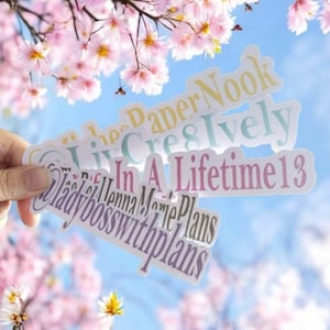 Peut inclure: Plusieurs autocollants avec du texte de différentes couleurs sont tenus par une main, sur fond de fleurs de cerisier roses et de ciel bleu. Les autocollants comportent des phrases telles que « Paper Nook », « Cres & Ively » et « In A Lifetime13 ».