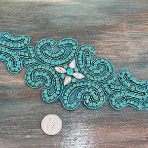 May include: An ornate, turquoise beaded applique with a central floral design and swirling patterns. The embellishment is approximately 6 inches long and features a mix of turquoise beads and silver accents. A coin is next to the applique for size comparison.