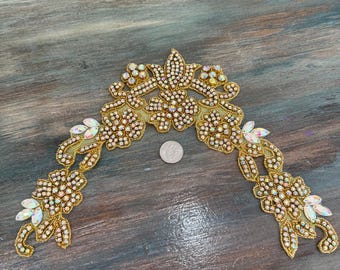 May include: A gold-colored, decorative applique with a floral design. The applique is embellished with iridescent rhinestones and small, clear crystals. The design is in an arch shape, with a coin for scale.