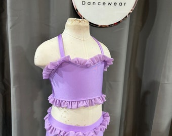 May include: A lavender dance leotard and matching shorts with a ruffled trim. The leotard has a halter style strap. The leotard and shorts are on a white mannequin.