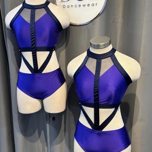 May include: Two dance costumes on mannequins. The costumes are a vibrant purple with black accents. Each has a halter top with mesh and a matching bottom. The costumes are displayed in front of a sign that says "DCD Dancewear".