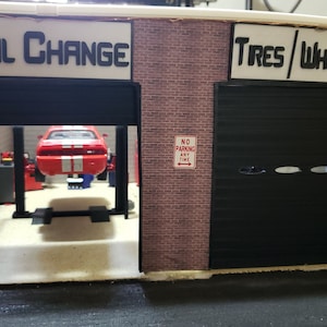 May include: A miniature model of a garage with three bays. The first bay is labeled "Oil Change" and has a red car on a lift. The second bay is labeled "Tires/Wheels" and the third bay is labeled "M". Each bay has a black garage door and a "No Parking Any Time" sign.