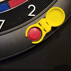 May include: Close-up of a yellow plastic dartboard accessory with a red button. The accessory is attached to a black dartboard with the number "17" and "2" visible.