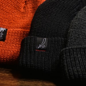 Puede incluir: Tres gorros de punto en naranja, negro y gris con un logotipo de palmera y ancla en cada uno.