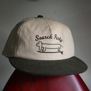 Może przedstawiać: Beżowa czapka z daszkiem z  cordu z zielonym daszkiem z cordu. Z przodu czapki znajduje się haftowany  dachshund na desce surfingowej z napisem "Search Party" nad nim.