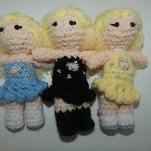 May include: Three crocheted dolls with blonde hair wearing different outfits. The doll on the left is wearing a light blue dress with a heart design. The doll in the middle is wearing a black dress with a sparkly design. The doll on the right is wearing a yellow dress with a heart design.