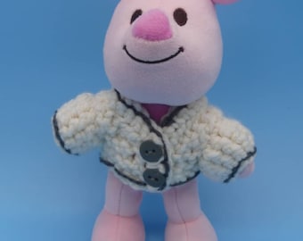 Könnte beinhalten: Ein rosa Plüschschwein, das einen weißen Häkelcardigan mit schwarzem Besatz und drei grauen Knöpfen trägt.