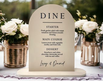 Könnte beinhalten: Ein bogenförmiges beiges Menüschild mit schwarzem Text, der DINE, STARTER, MAIN COURSE, DESSERT und die Namen Lucy & David zeigt. Das Schild ist auf einem flachen ovalen Sockel montiert und enthält eine detaillierte Liste von Speisen wie Salat und Forelle.