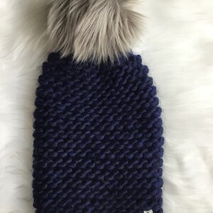 May include: A navy blue knitted beanie with a gray faux fur pom pom. The beanie has a small label that says "Lori's Knits".