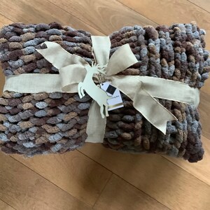 May include: A brown, gray, and beige knitted throw blanket with a beige ribbon tied in a bow. The blanket is decorated with a white wooden reindeer ornament and a tag that reads "Cozy Knit".