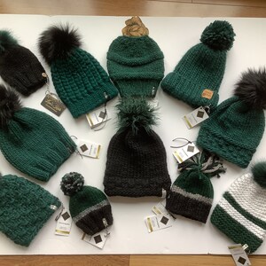 May include: Assortment of green and black knit hats with faux fur pom poms. Some hats have a white stripe.