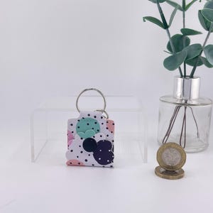 May include: A white keychain with a colourful polka dot pattern and Mickey Mouse-shaped cutouts in mint green, pink, and navy blue. The keychain has a silver ring and is displayed on a clear acrylic stand.