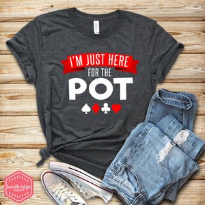 May include: A dark grey t-shirt with the text "I'm just here for the POT" in white with a red ribbon above the text.  Four playing card suits are below the text: a spade, a diamond, a club, and a heart.