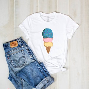 Peut inclure: T-shirt blanc avec un motif de cornet de glace &agrave; trois boules avec des p&eacute;pites sur le dessus. La glace est bleue, rose et jaune. Le cornet est brun. Le t-shirt est pos&eacute; sur une surface en bois avec un jean bleu.