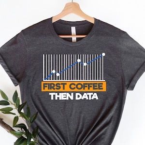 Puede incluir: Una camiseta gris oscuro con un gráfico de líneas blancas y una línea azul. El gráfico muestra una tendencia al alza. El texto "First Coffee Then Data" está impreso en naranja en la camiseta.