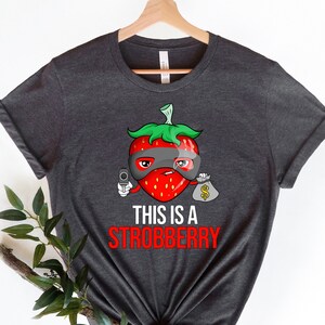 May include: A dark gray t-shirt with a cartoon strawberry wearing a mask and holding a gun and a bag of money. The text "This is a Strobbberry" is printed below the strawberry.