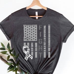Puede incluir: Una camiseta gris oscuro con un diseño de bandera estadounidense desgastada y una cinta de estampado de cebra. El texto "FIGHT" está impreso verticalmente en la bandera.