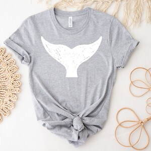 May include: A gray t-shirt with a white whale tail graphic. The shirt is tied in a knot at the bottom.