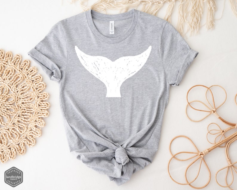 May include: A gray t-shirt with a white whale tail graphic. The shirt is tied in a knot at the bottom.