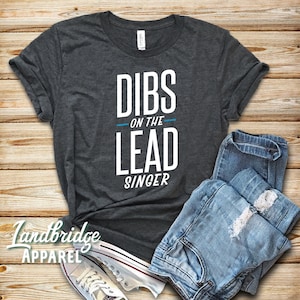 May include: A dark grey t-shirt with the text "DIBS ON THE LEAD SINGER" in white letters. The text "ON THE" is in blue.