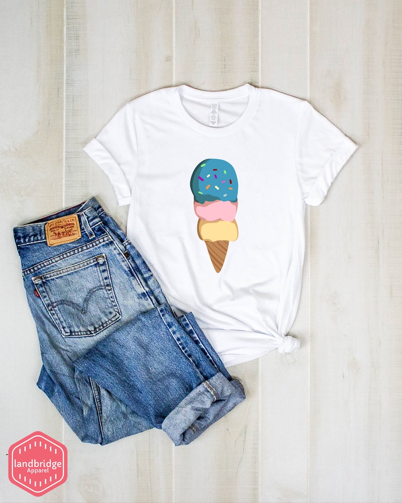 Peut inclure: T-shirt blanc avec un motif de cornet de glace &agrave; trois boules avec des p&eacute;pites sur le dessus. La glace est bleue, rose et jaune. Le cornet est brun. Le t-shirt est pos&eacute; sur une surface en bois avec un jean bleu.