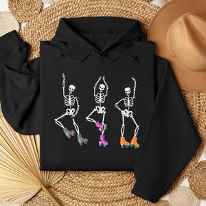K&ouml;nnte beinhalten: Schwarzer Hoodie mit einer wei&szlig;en Grafik von drei Skeletten, die Rollschuh fahren. Die Skelette tragen Rollschuhe mit orangefarbenen und rosafarbenen Rollen.