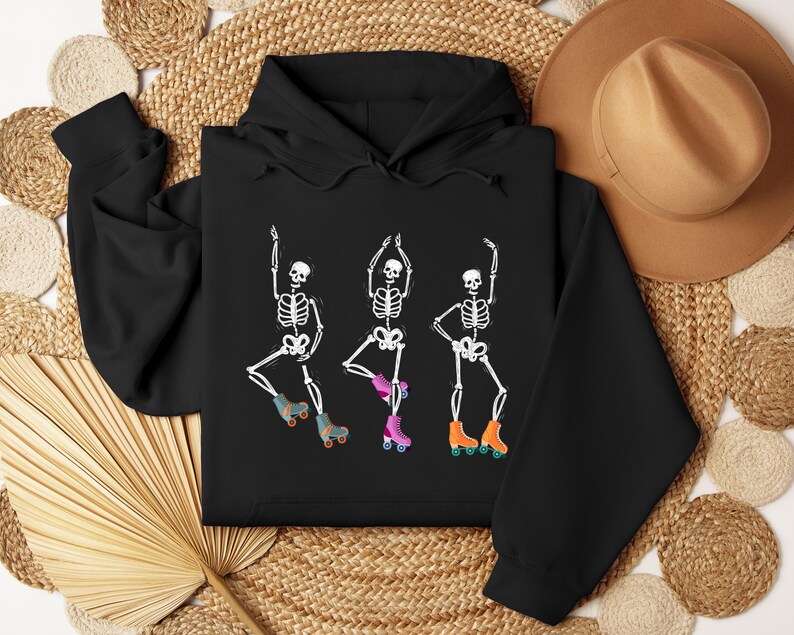 K&ouml;nnte beinhalten: Schwarzer Hoodie mit einer wei&szlig;en Grafik von drei Skeletten, die Rollschuh fahren. Die Skelette tragen Rollschuhe mit orangefarbenen und rosafarbenen Rollen.