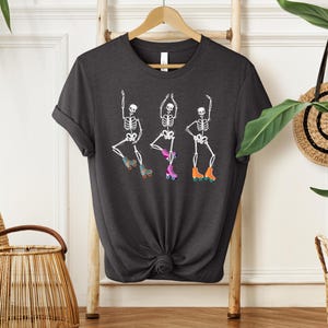 K&ouml;nnte beinhalten: Ein dunkelgraues T-Shirt mit einer wei&szlig;en Grafik von drei Skeletten, die Rollschuh laufen. Die Skelette tragen bunte Rollschuhe.