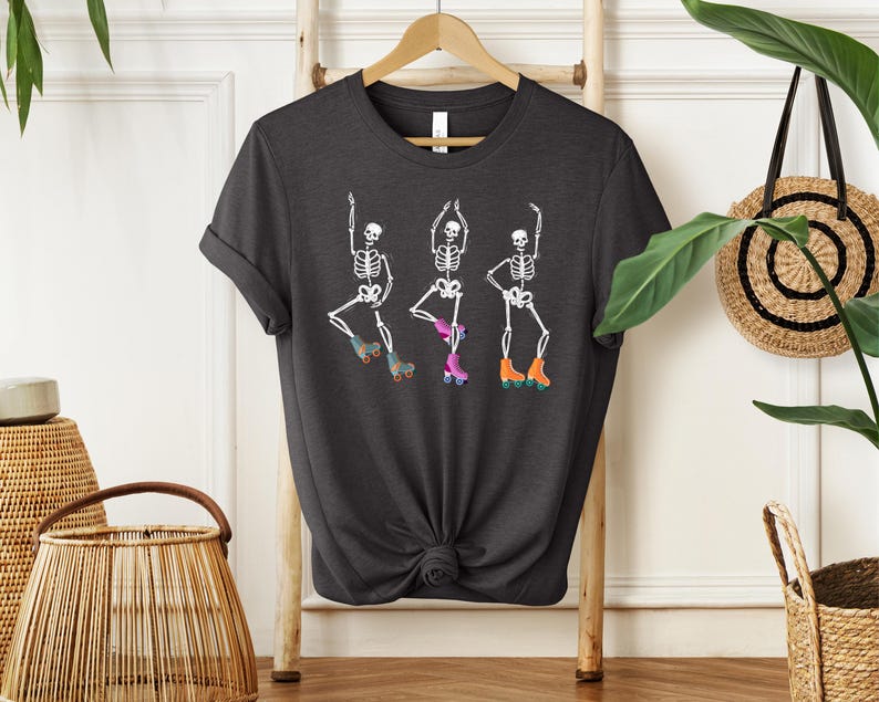 K&ouml;nnte beinhalten: Ein dunkelgraues T-Shirt mit einer wei&szlig;en Grafik von drei Skeletten, die Rollschuh laufen. Die Skelette tragen bunte Rollschuhe.