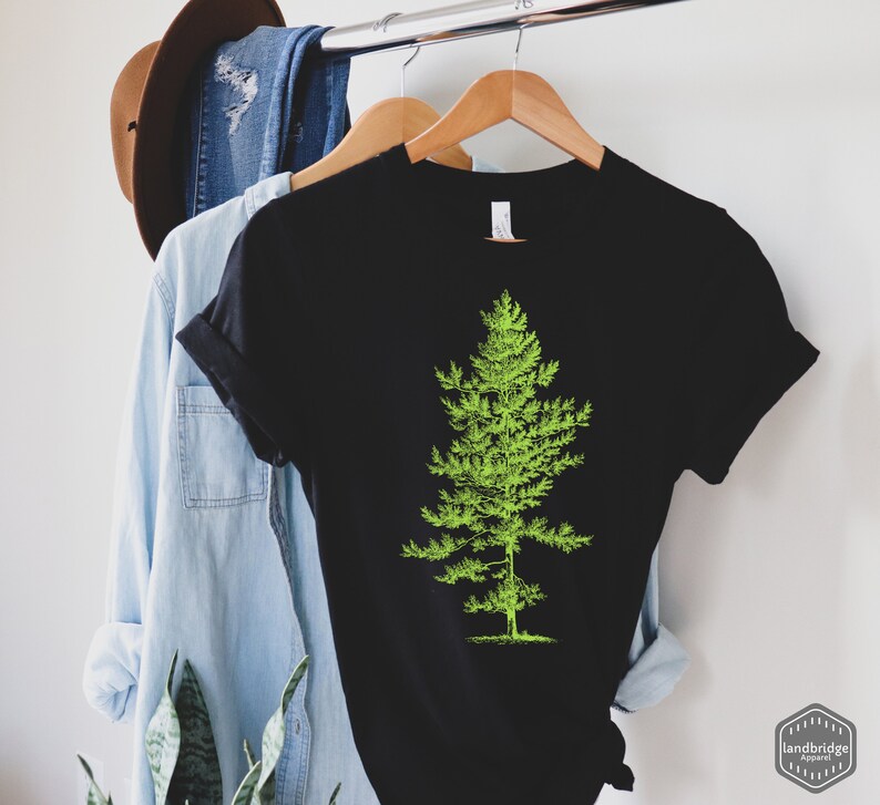 May include: Black t-shirt with a green silhouette of a pine tree on the front. The shirt is hanging on a hanger.