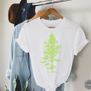 May include: White t-shirt with a green graphic of a pine tree. The t-shirt is hanging on a wooden hanger.