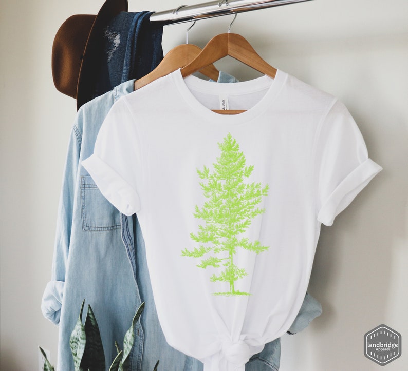 May include: White t-shirt with a green graphic of a pine tree. The t-shirt is hanging on a wooden hanger.