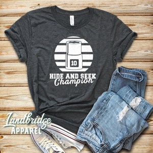 May include: A dark grey t-shirt with a white graphic of a spanner with the number 10 on it. The spanner is inside a white circle with white stripes. The text "Hide and Seek Champion" is below the circle.