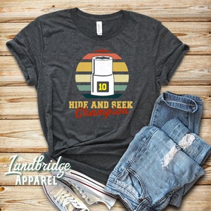 Könnte beinhalten: Ein dunkelgraues T-Shirt mit einem Vintage-Sonnenuntergang-Motiv, das einen Schraubenschlüssel mit der Nummer 10 zeigt. Der Text "Hide and Seek Champion" ist unter dem Motiv gedruckt.