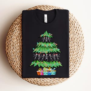 Op de afbeelding: Zwart T-shirt met een groene kerstboom versierd met skeletten en lampjes. De boom is omringd door kleurrijke ingepakte cadeaus.