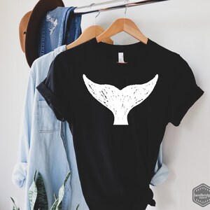 May include: Black t-shirt with a white whale tail graphic. The t-shirt is hanging on a wooden hanger.