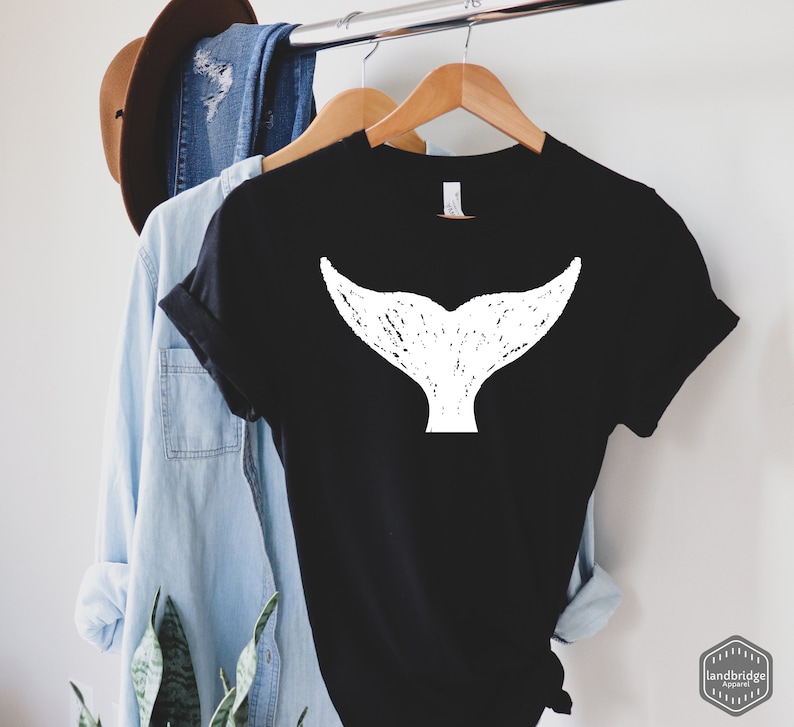 May include: Black t-shirt with a white whale tail graphic. The t-shirt is hanging on a wooden hanger.
