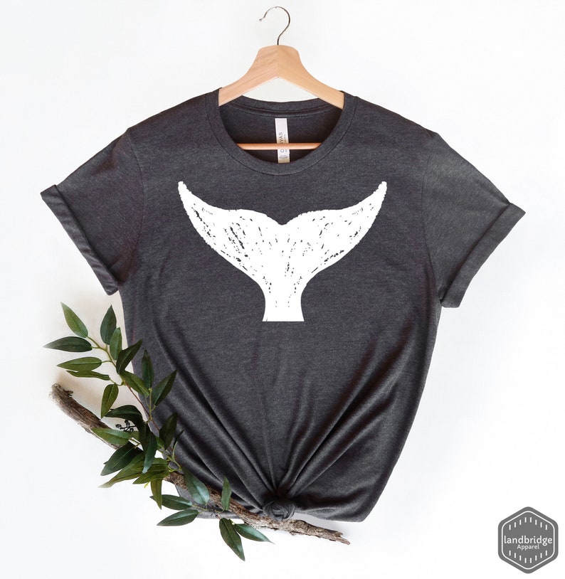 May include: A dark gray t-shirt with a white whale tail graphic. The tail is stylized with a distressed look. The shirt is on a hanger.