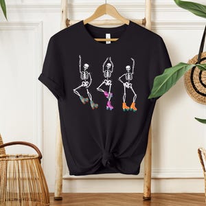 K&ouml;nnte beinhalten: Schwarzes T-Shirt mit einem Motiv von drei Skeletten, die Rollschuh laufen. Die Skelette tragen bunte Rollschuhe und sind in verschiedenen Posen.