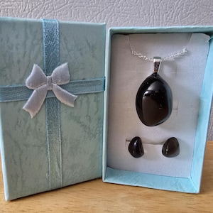 May include: A jewellery set in a pale blue gift box, tied with a silver ribbon and bow. Inside, a black pendant necklace and matching stud earrings are presented on a light blue insert. The pendant and earrings have a polished, glossy finish.