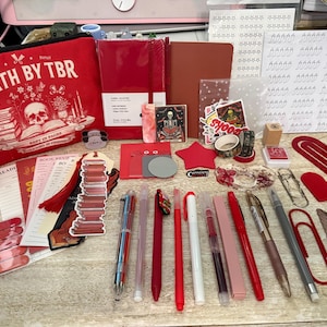 May include: A collection of stationery and reading accessories including a red pouch with the text DEATH BY TBR, various red and neutral-toned notebooks, pens, stickers, paper clips, a bookmark shaped like a stack of books, and a feather-shaped item.