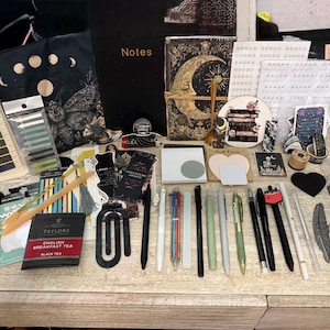 May include: A collection of stationery items including a black notebook labeled Notes, a journal with a gold moon illustration, various pens, markers, sticky notes, bookmarks, paper clips, stickers, a tea bag, and decorative paper ephemera.